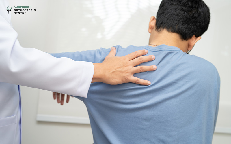 Medical professional in white coat examining a seated patient's shoulder in a clinical setting.