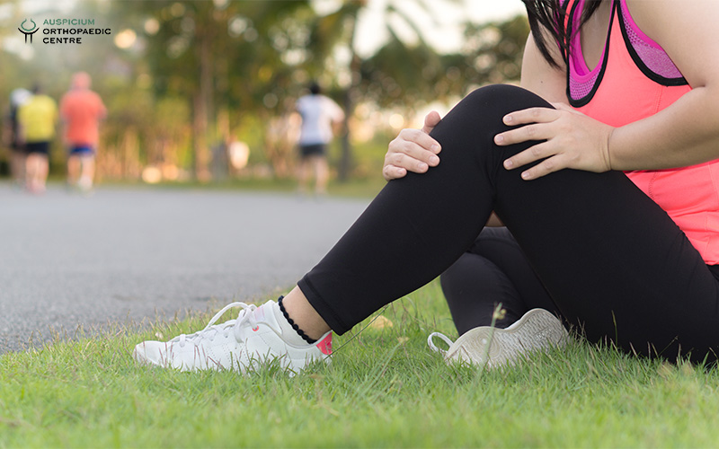 Person in pink athletic top sitting on grass beside a path, holding knee in pain at a park.