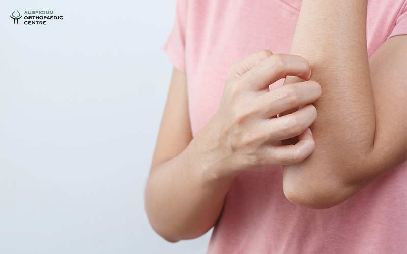 Person in pink shirt scratching left forearm, suggesting skin irritation, against a plain light background.