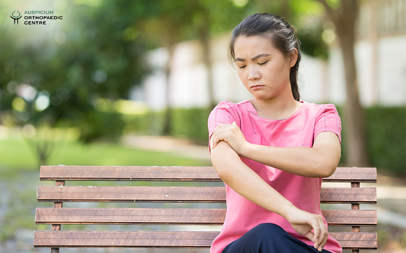 Person in pink shirt sitting on a bench outdoors, holding left arm in discomfort near greenery and buildings.