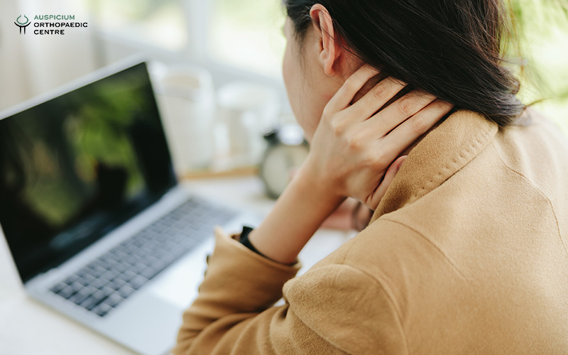 Person in tan jacket seated at desk holding neck in pain, facing a laptop with dark screen.