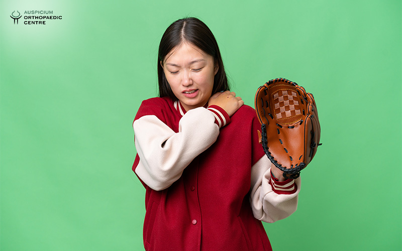 Person in varsity jacket holding a baseball glove and touching shoulder, indicating sports-related shoulder pain.