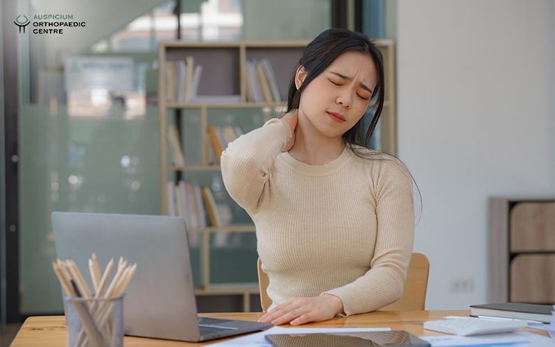 Person seated at a desk holding neck in pain, with laptop and documents in a modern office setting.