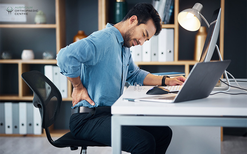 Man in office setting leaning forward at desk, supporting lower back with hand while working on laptop.