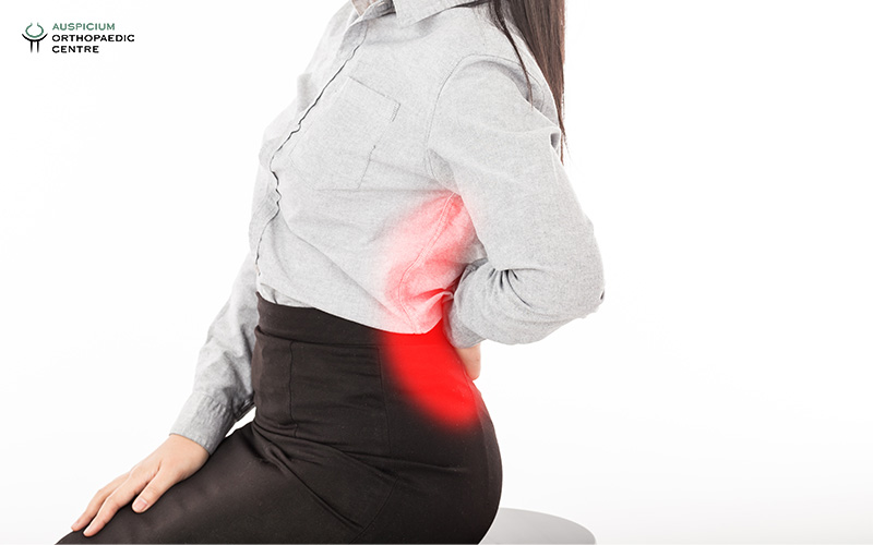 Person in gray shirt seated on stool holding lower back in pain, with red glow indicating discomfort.